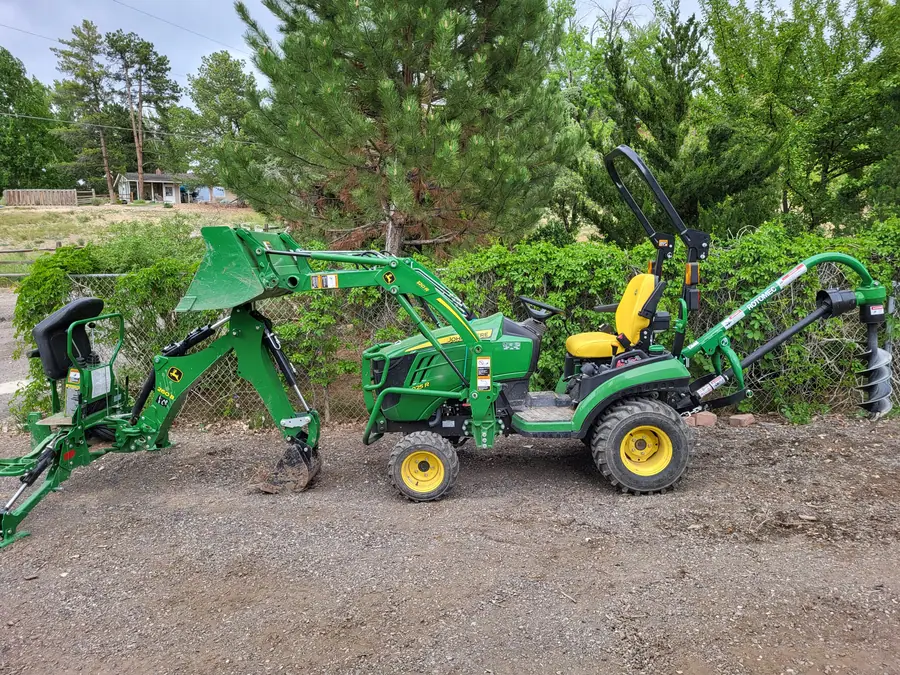 A green compact tractor with a front loader, a backhoe and a post-hole auger, parked on gravel in front of a hedge.